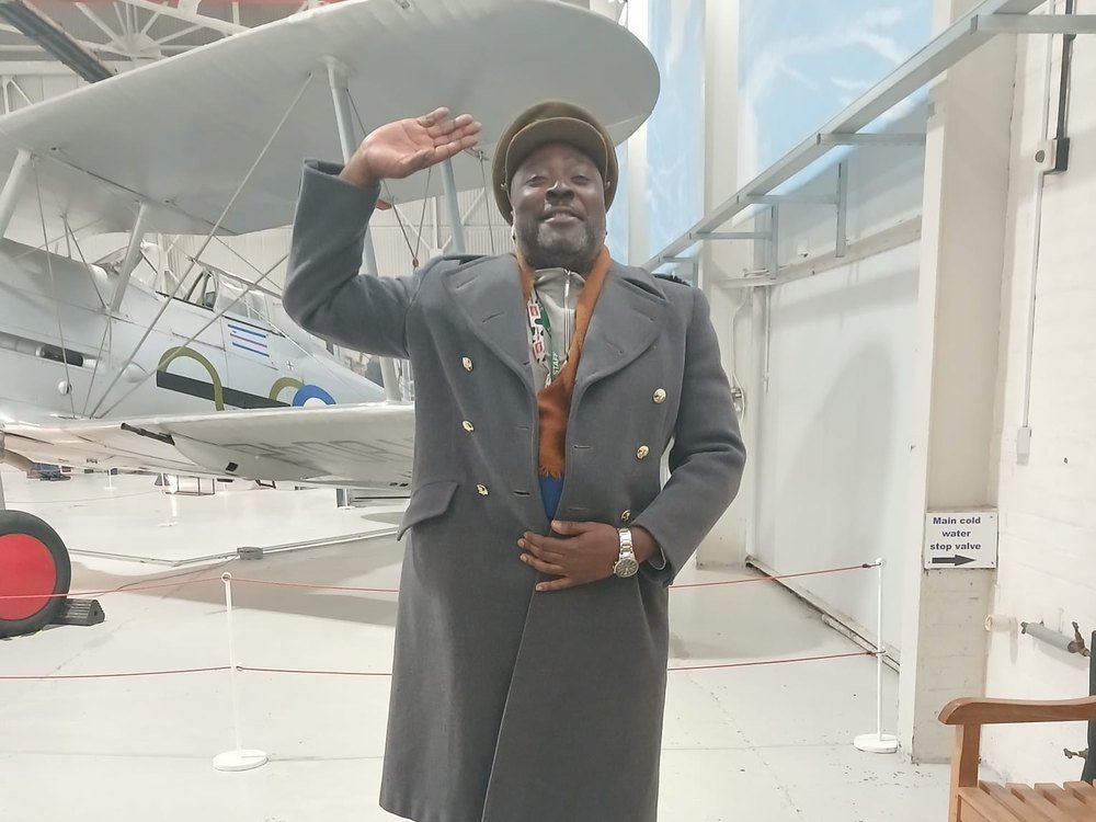 A member of staff in a period greatcoat and cap saluting beside a biplane at the RAF museum