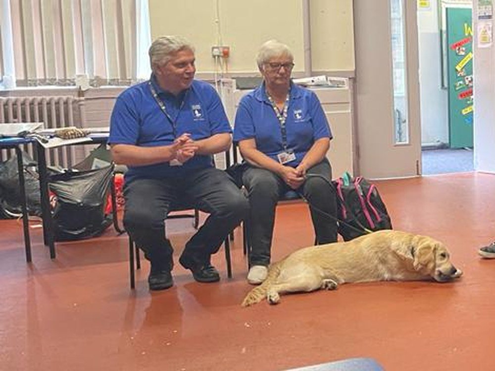 Two visitors from a guide dog charity sitting with a guide dog during a school visit